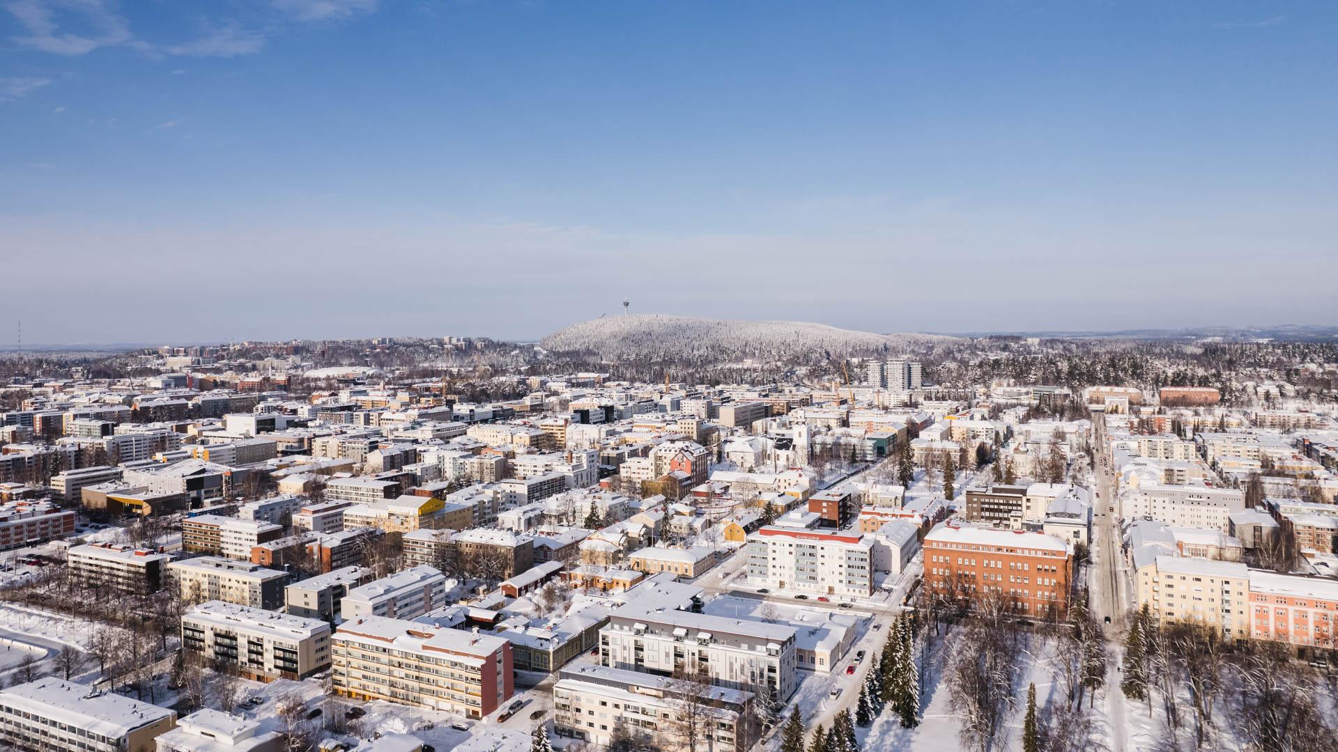 Ilmakuva talvisesta Kuopion keskustasta, taustalla lumipeitteinen Puijo ja Puijon torni.