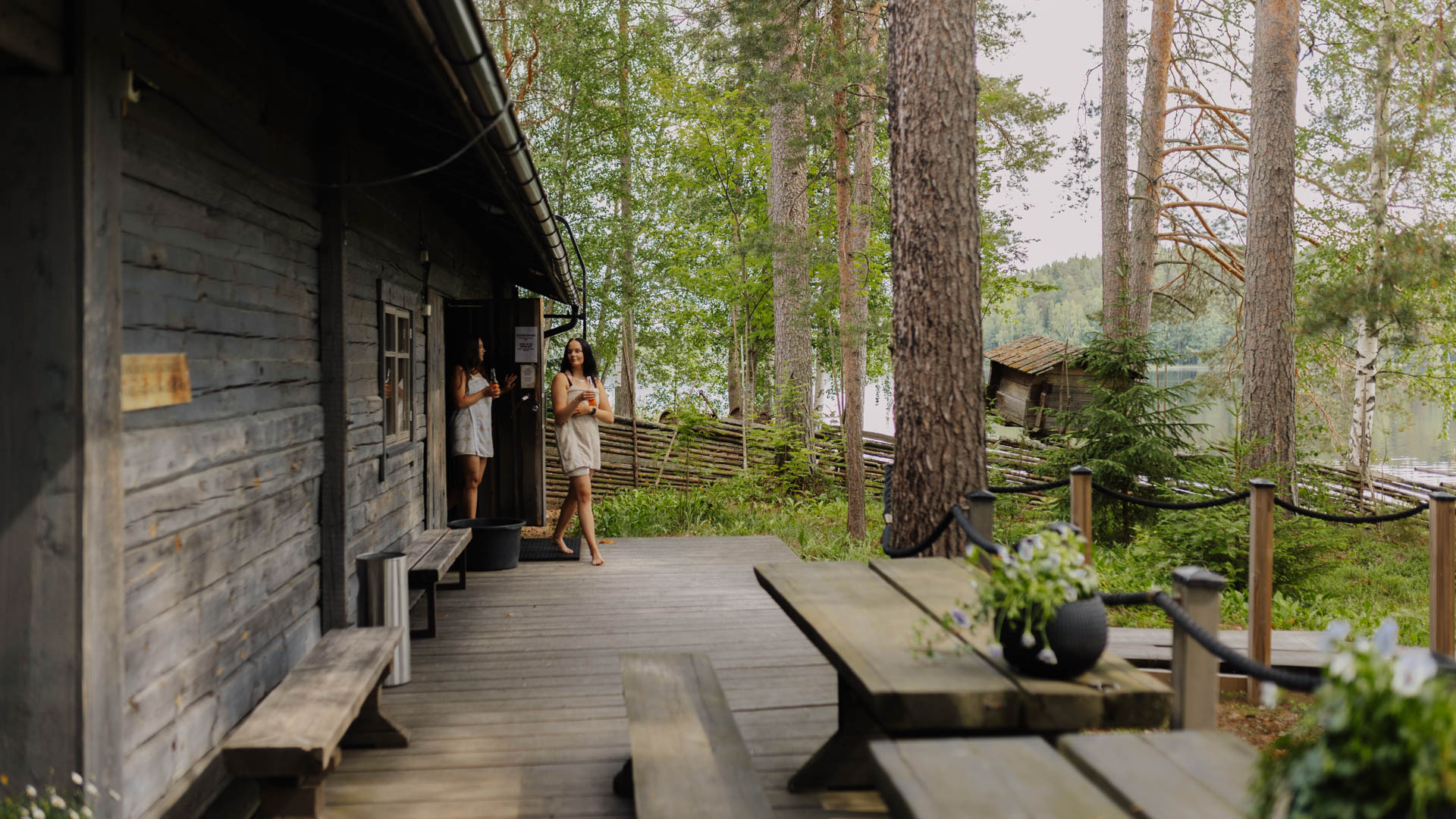Rauhalahti-Jatkankamppa-perinne-ja-savusaunailta Ihmisiä rentoutumassa perinteisen savusaunan terassilla metsä- ja järvimaisemissa.